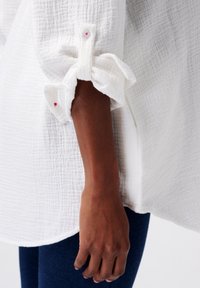 Un bras à la peau foncée, partiellement recouvert par une chemise à manches longues blanches texturées retroussée, sur un fond blanc avec un pantalon bleu marine.