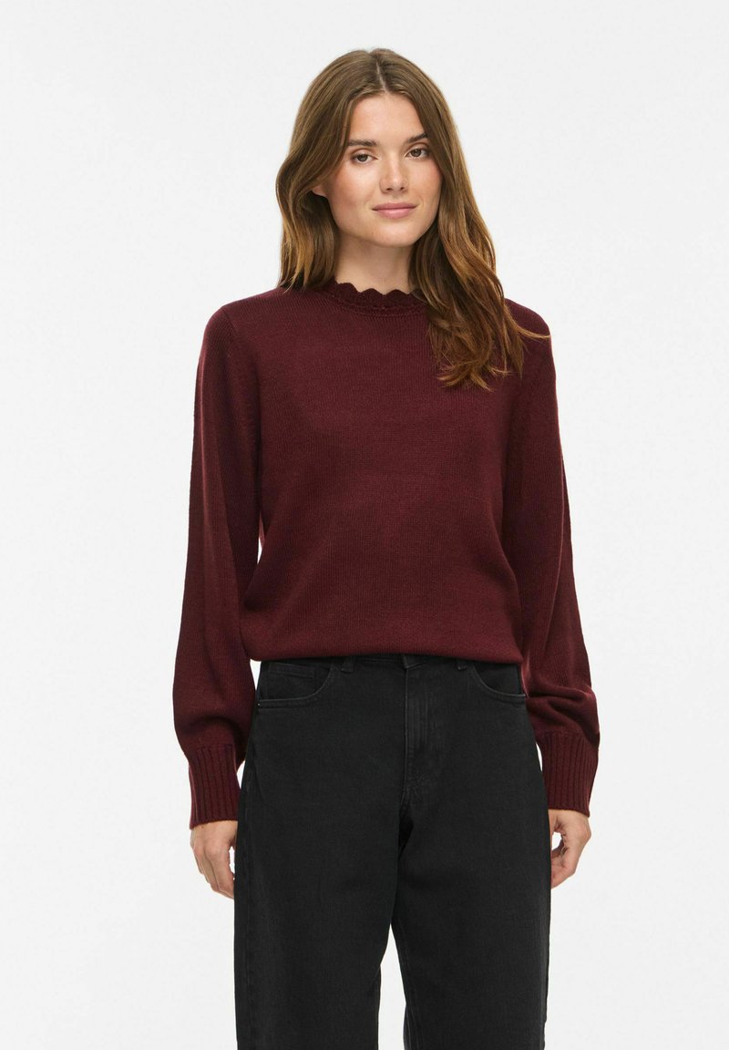 Femme aux longs cheveux bruns portant un pull bordeaux à col festonné et un pantalon noir taille haute sur fond blanc.