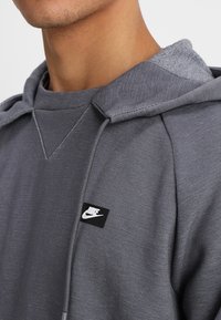 Grå hoodie i ett texturerat tyg, med en känguruficka, dragsko i huvan och en liten svart Nike-logotyp på framsidan.