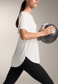 Top sportiv scurt alb cu mâneci scurte și hem rotunjit, purtat cu colanți negri. Persoana ține în mână o minge de exerciții gri texturată.
