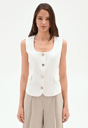 Donna che indossa un gilet bianco senza maniche con bottoni e tasche con pattina e pantaloni beige a righe sottili a gamba larga su sfondo bianco.