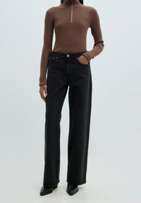Zwarte high-waisted wijde jeans met een knoop- en ritsluiting, gecombineerd met een geribde bruine coltrui en zwarte puntige laarzen.