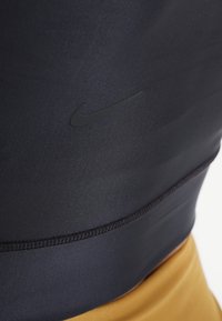 Schwarzes Sportoberteil mit einer weichen, glatten Textur, das mit einem kleinen schwarzen Nike-Logo und Nähten versehen ist, kombiniert mit golden gefärbtem Stoff.