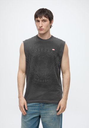 Jeune homme aux cheveux courts et foncés, portant un t-shirt noir sans manches Diesel et un jean bleu clair, faisant face à un fond uni.