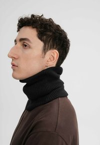 Zwarte geribbelde coltrui met een hoge kraag. Model draagt een bruine blouse, met korte krullen en een kleine oorbel zichtbaar.