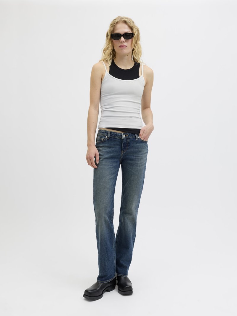 Vit linne över en svart topp, blå denimjeans och svarta klumpiga skor. Långt vågigt hår och mörka solglasögon.