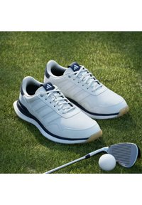Chaussures de golf Adidas blanches sur l'herbe à côté d'un club de golf et d'une balle de golf blanche.