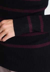 Black ribbed sweater featuring maroon stripes along the sleeves. Close-up of the texture and cuff design with a fitted shape.