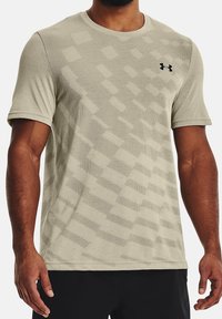 Man med en beige kortärmad träningströja med ett geometriskt mönster och svart Under Armour-logotyp på bröstet.