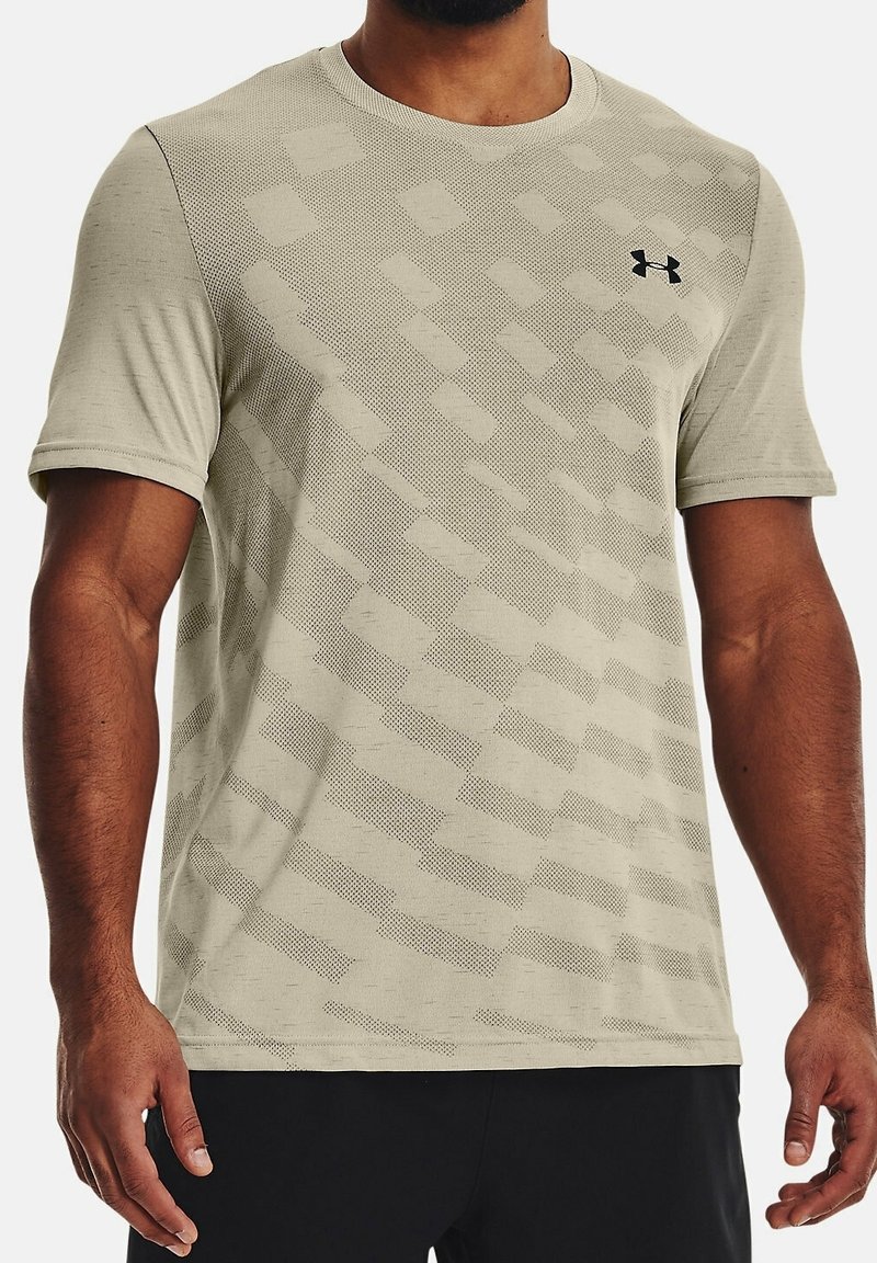 Man med en beige kortärmad träningströja med ett geometriskt mönster och svart Under Armour-logotyp på bröstet.