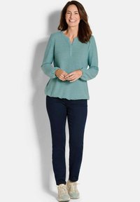 Maglione verde acqua a maniche lunghe con scollatura a henley e polsini arricciati, abbinato a pantaloni scuri slim-fit e sneaker chiare.
