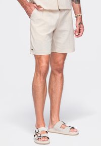 Homme portant un short beige avec des poches et des sandales beige clair à enfiler avec des boucles noires, debout sur un fond blanc.