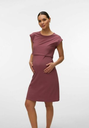 Gravid kvinde står, iført en tætsiddende, ærmeløs lyserød graviditetskjole, der ømt holder sin mave med begge hænder.