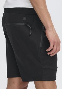 Svarta shorts i mjukt tyg, med en åtsittande midja, dragkedjefickor på sidan och en cirkulär logotypdetalj på baksidan.