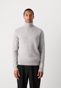 Pull à col roulé gris clair en tissu doux, avec des poignets et un ourlet côtelés. Positionné sur un fond uni de ton neutre.