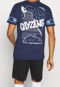 Man som bär en marinblå Puma-t-shirt med ett fragmenterat mönster och stor text "CITIZENS" över upprepade "MANCHESTER" på ryggen, svarta shorts, vita strumpor.
