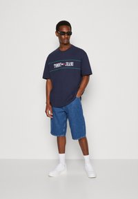 T-shirt in cotone blu navy con logo "TOMMY JEANS" in bianco e rosso, abbinato a pantaloni shorts in denim chiaro, sneakers bianche e occhiali da sole neri.