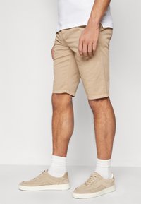 Mann in beige Knielangen Shorts, beigen Sneakers mit weißen Sohlen und weißen Sneakersocken, steht vor einem schlichten weißen Hintergrund.