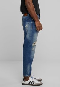 Jeans de denim azul con un ajuste relajado, que presentan detalles desgastados y zonas desvanecidas. Combinados con zapatillas blancas y una camiseta negra.