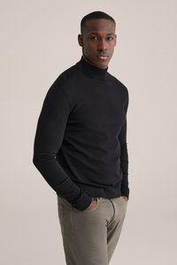 Melna augsta apkakle džemperis no gludas auduma, piegulošs dizains, garas piedurknes, valkāts kopā ar gaiši pelēkām biksēm, stāvot pret neitrālu fonu.