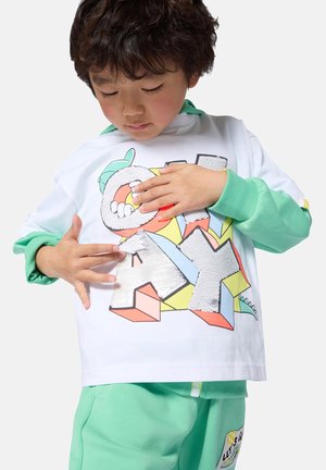 Enfant portant un pantalon vert et un sweat-shirt blanc avec un graphisme coloré "PLAY", touchant les lettres en sequins sur le haut.
