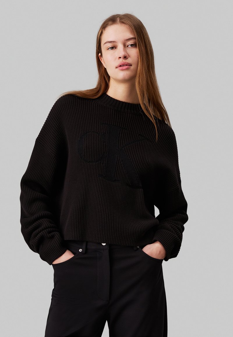 Maglione lavorato a maglia nero con una texture a coste, scollo rotondo e un grande logo "CK" in una tonalità abbinata. Abbinato a pantaloni neri.