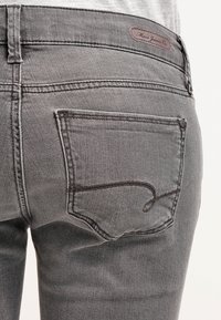 Jeans in denim grigio con una tasca posteriore caratterizzata da un design di cucitura curva e un patch in pelle sulla vita. Texture liscia.