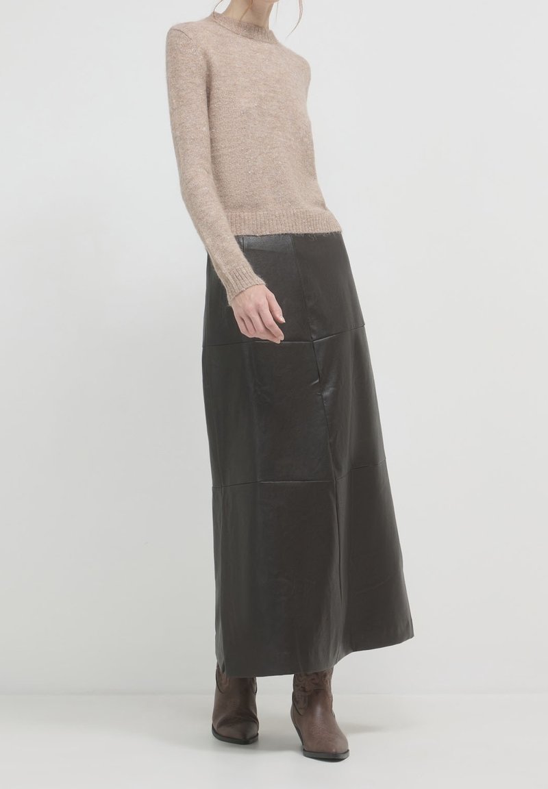 Femme portant un pull en maille beige, longue jupe en cuir noire et bottines marron, debout devant un fond blanc uni.