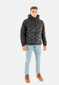 Chaqueta acolchada negra con cremallera y capucha, con un frente texturizado. Se lleva sobre unos vaqueros azul claro y calzado marrón.