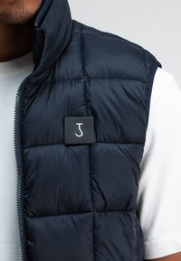 Marineblauer gesteppter Weste mit hohem Kragen, ausgestattet mit einem schwarzen quadratischen Logo-Patch und einem durchgehenden Reißverschluss, getragen über einem hellgrauen T-Shirt.