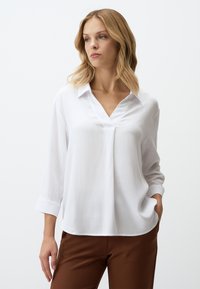 Blouse blanche à manches longues avec un col en V et un tissu texturé. Dotée d'un col et de manches à poignets. Associée à un pantalon marron.