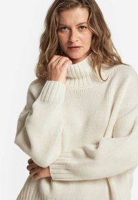 Flauschiger Strickpullover in Cremefarbe mit hohem Rollkragen, gerippten Bündchen und lockerer Passform. Die strukturierte Webart verleiht dem Pullover Dimension.