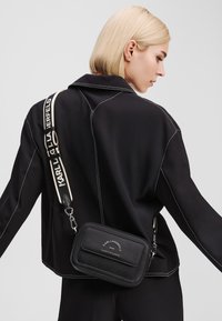 Black textured crossbody bag with adjustable strap. Features silver hardware and logo detailing. Background is plain gray.