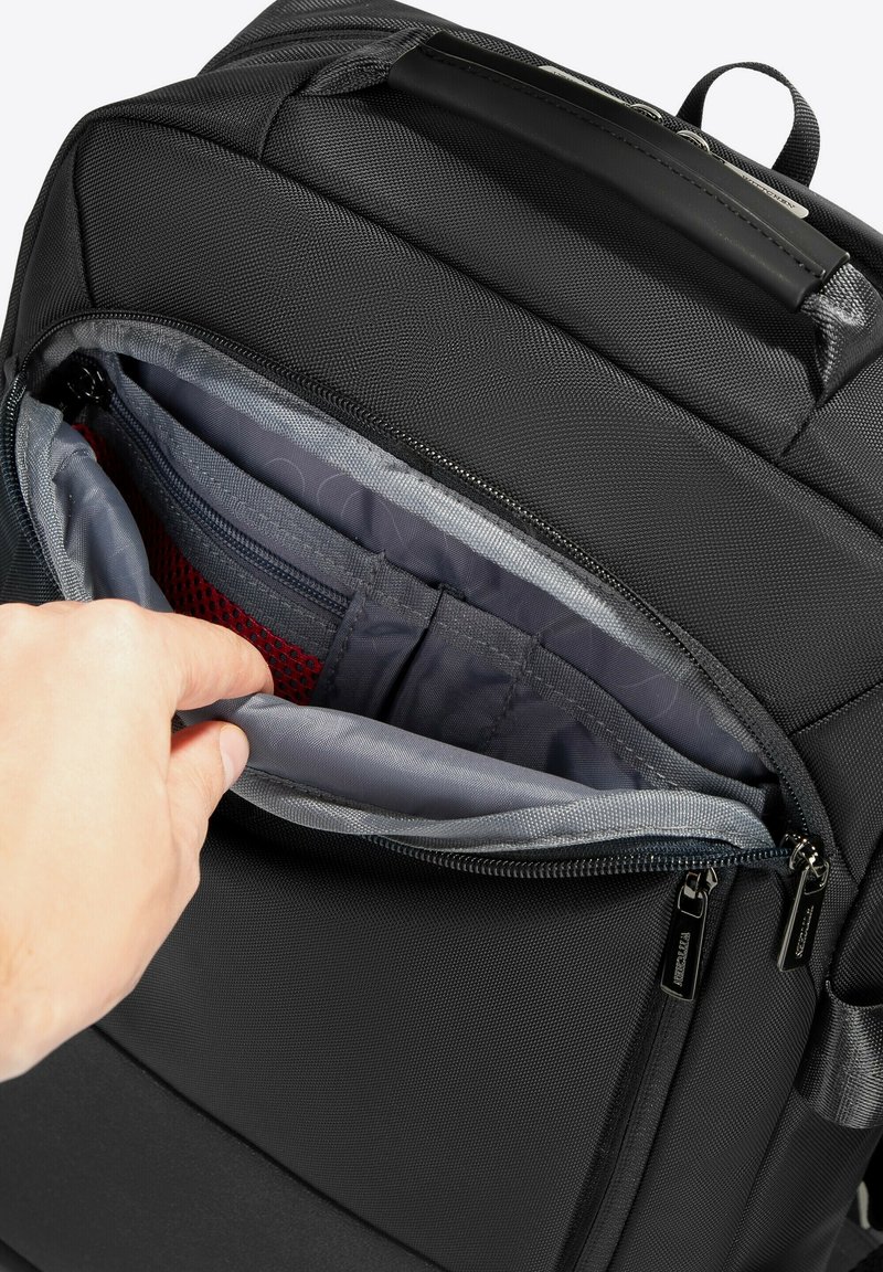 Sac à dos en tissu noir avec une poche zippée ouverte, révélant des compartiments intérieurs gris et rouge et une disposition organisée pour les accessoires.