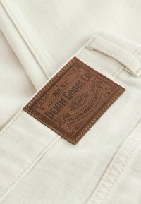 Beige tyg med en sydd brun läderetikett som läser "NEXT DENIM GOODS CO" och ornamentala detaljer, satt mot en strukturerad bakgrund.