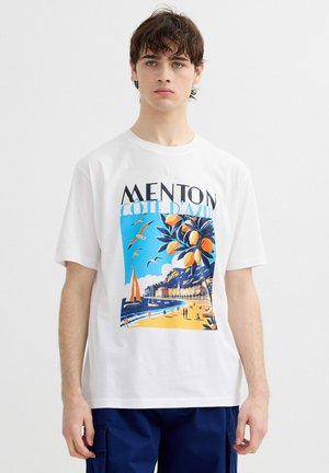 Joven con una camiseta blanca con un dibujo colorido de la playa de Menton Côte d'Azur, gaviotas, veleros y árboles de naranja.