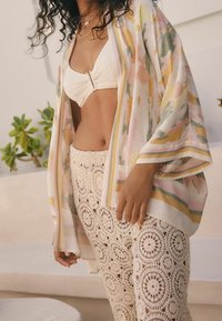 Letthåndterlig, flerfarvet kimono med abstrakte mønstre, parret med en struktureret off-white hæklet nederdel. Blødt stof med flagrende ærmer.