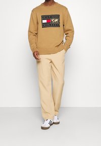 Maglione beige lavorato a maglia con logo grafico, abbinato a pantaloni a gamba larga beige chiaro e sneakers bianche con strisce nere.