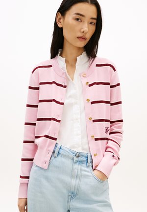 Jonge vrouw die een roze en kastanjebruine gestreepte cardigan draagt, een witte blouse en lichtblauwe spijkerbroek, met één hand in haar zak.