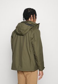 La giacca impermeabile verde oliva presenta un cappuccio, chiusura con zip e polsini elasticizzati, con un design pulito e minimalista senza loghi visibili.