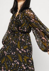 Černé šaty s květinovými a abstraktními vzory v růžové, zelené a bílé. Obsahují dlouhé, průsvitné rukávy a detail se zpevněným pasem.