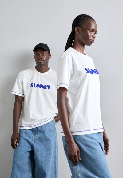 Λευκά μπλουζάκια με μπλε κεντητό λογότυπο "SUNNEI", σε συνδυασμό με ανοιχτόχρωμες παντελόνες σε φαρδιά γραμμή. Ένα μοντέλο φοράει ένα μαύρο καπέλο και έχει υφασμάνα πλεγμένα μαλλιά.