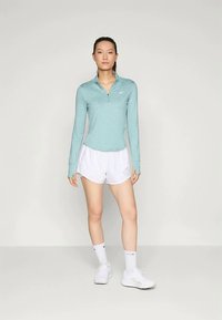 Frau steht in einem hellblauen Langarm-Sportoberteil, weißen Shorts, weißen Socken und weißen Turnschuhen vor einem schlichten Hintergrund.