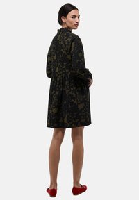 Robe à motifs noirs et olive avec des manches longues, un col volanté et une taille froncée. Portée avec des ballerines rouges.