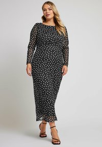 Musta läiskäinen mekko, jossa on pitkät läpinäkyvät hihat, istuva vartalo-osa ja valtaisa hame. Mekossa on pyöreä pääntie ja nilkkapituinen helma.