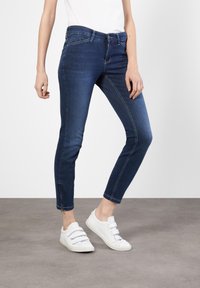 Dunkelblaue Denim-Jeans mit slim fit, mit einem mittelhohen Bund, kontrastierenden Nähten und Reißverschlüssen an den Knöcheln. Dazu weiße Sneakers.