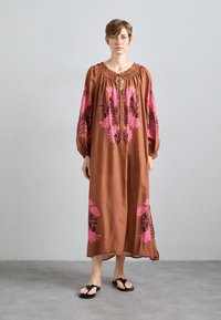 Braun, fließendes Kleid mit großen pinken Blumenmustern, runder Halsauschnitt und gerafftem Detail. Lange Ärmel verjüngen sich an den Handgelenken. Getragen mit schwarzen Sandalen.