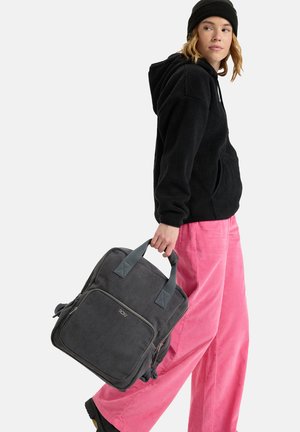 Grijze corduroy rugzak met dubbele handgrepen bovenaan, een voorvak en een ritsluiting, gedragen door een persoon in een zwarte fleece hoodie en roze broek.