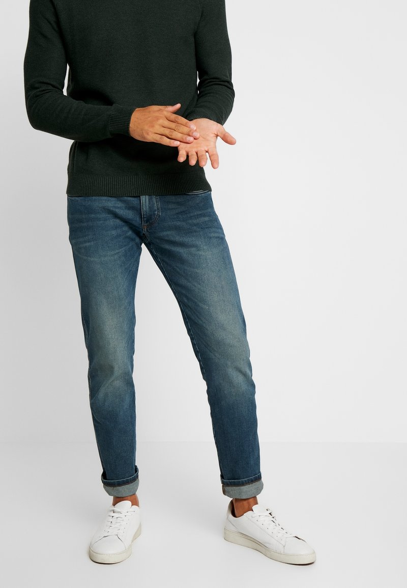 Dunkelgrüner gestrickter Pullover kombiniert mit hellblauen Jeans, hochgekrempelt an den Manschetten, und weißen Sneakers. Die Hände gestikulieren. Weißer Hintergrund.