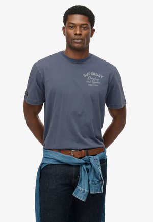 Mørkeblå bomulds t-shirt med rund hals, med hvid "SUPERDRY" logo. Båret med mørke jeans og en lyseblå denimskjorte knyttet om taljen.